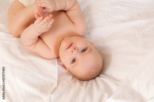 Cute baby in diapers lies in bed with white cotton linen, view from above, space for text