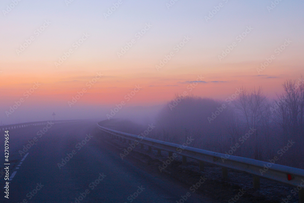 A bright dawn in pastel colors and with heavy fog greets us on a country road.