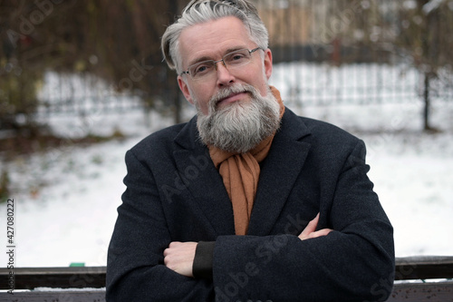 Serious adult gray-haired man with glasses sits on a bench and looks at the camera