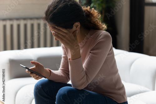 Out of the blue. Frustrated millennial latin woman sit on sofa read sudden bad news from cell screen cover face with palm in desperate gesture. Stressed young lady get nasty blackmail message on email photo