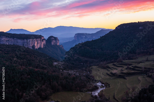 Landscape of a valley with towering rocky mountains with a beautiful sky with pink and orange clouds at sunset. Cingles de Tavertet, Collsacabra, Osona, Barcelona, Catalonia, Spain.