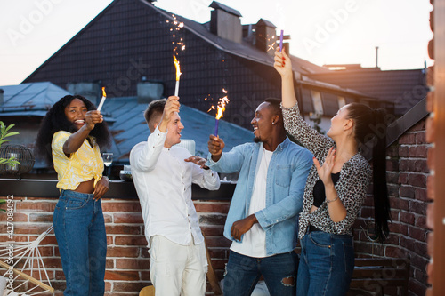 Joyful multiracial friends smiling, chatting and using sparklers during rooftop party. Young hipster people enjoying leisure time spending together.