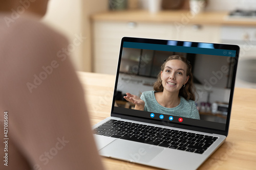 Smiling young woman call sister mother using laptop video app talk share good news. Pleasant millennial lady on laptop screen hold online dialogue with remote friend at webcam chat. Over shoulder view