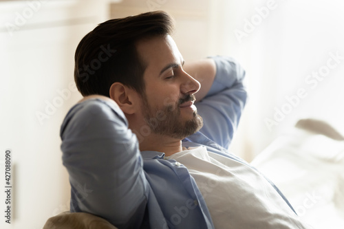 Young Caucasian man sit relax on couch breathe fresh ventilated condition air at home. Calm millennial male rest on sofa sleep or take nap, relieve negative emotions. Stress free concept.