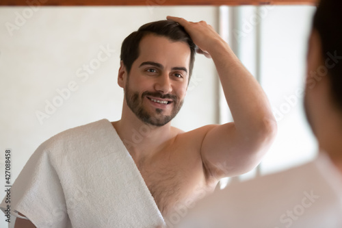 Картината върху платно Happy millennial handsome man in towel look in mirror in bathroom at home get ready in morning