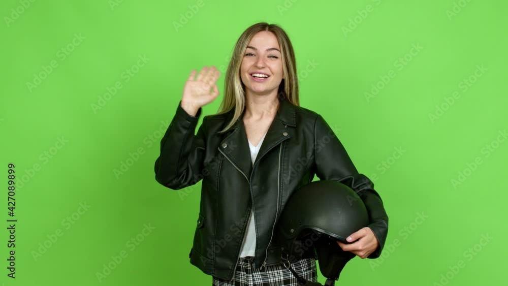 Young woman holding a motorcycle helmet saluting with hand with happy expression over isolated background on green screen chroma key Young woman holding a motorcycle helmet saluting with hand with happy expression over isolated background on green screen chroma key