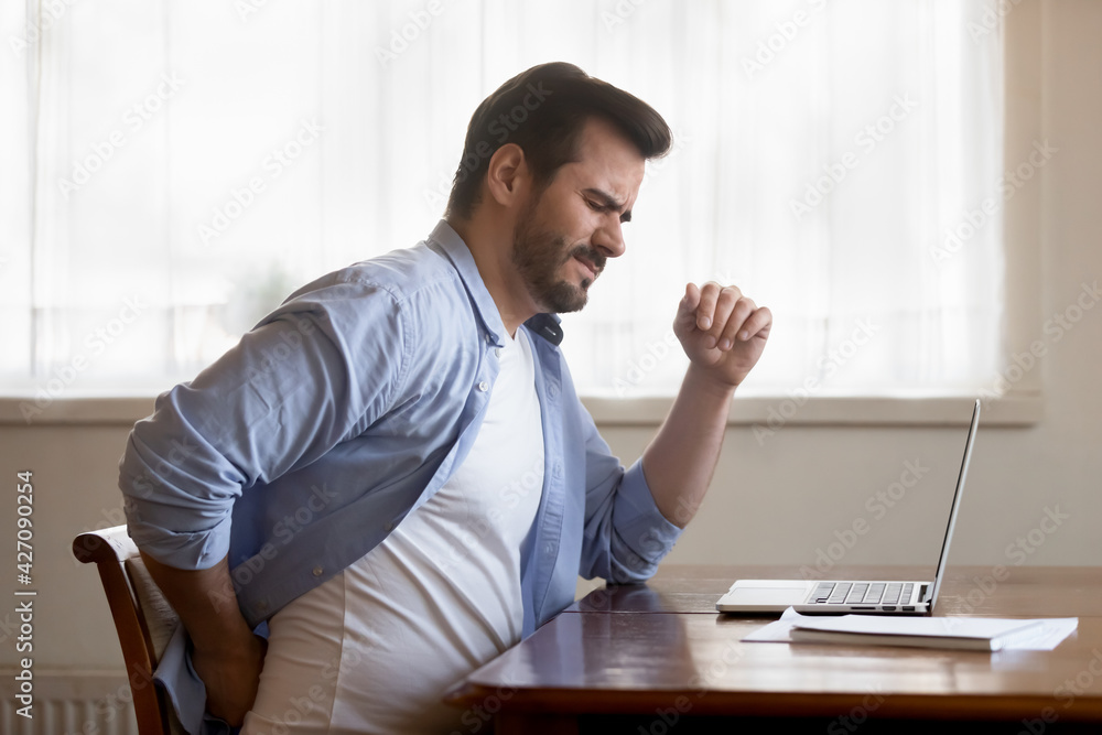Unhappy Caucasian man sit at desk in home office work on laptop suffer from lower back muscle strain or spasm. Unwell unhealthy male have backache from incorrect posture. Sedentary life concept. Unhappy Caucasian man sit at desk in home office work on laptop suffer from lower back muscle strain or spasm. Unwell unhealthy male have backache from incorrect posture. Sedentary life concept.