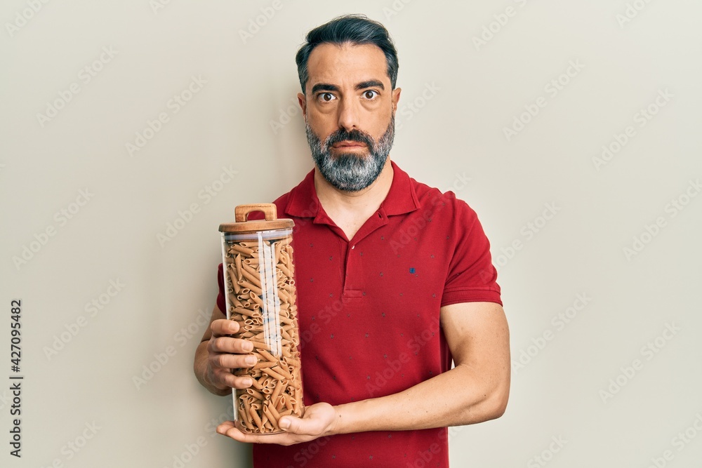 Fototapeta premium Middle age man with beard and grey hair holding jar with macaroni pasta in shock face, looking skeptical and sarcastic, surprised with open mouth