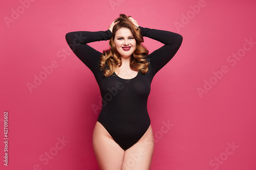 A young plus-size model woman with bright makeup and full red lips wearing a black bodysuit isolated over pink background photo