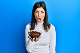 Young hispanic woman holding raisins bowl scared and amazed with open mouth for surprise, disbelief face