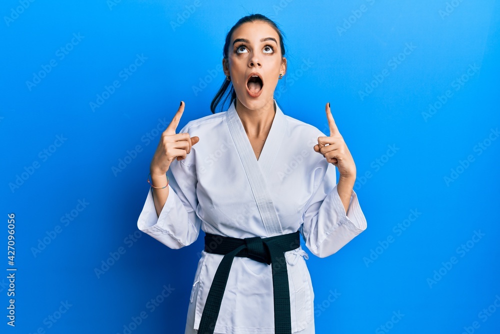 Beautiful brunette young woman wearing karate fighter uniform with black belt amazed and surprised looking up and pointing with fingers and raised arms. Beautiful brunette young woman wearing karate fighter uniform with black belt amazed and surprised looking up and pointing with fingers and raised arms.