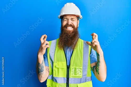 Redhead man with long beard wearing safety helmet and reflective jacket gesturing finger crossed smiling with hope and eyes closed. luck and superstitious concept.