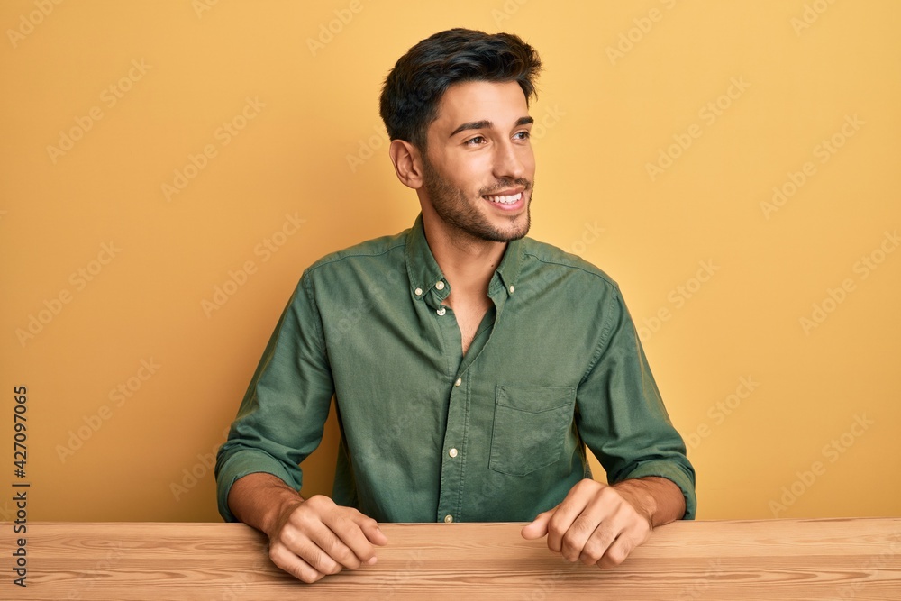 Young handsome man wearing casual clothes sitting on the table looking to side, relax profile pose with natural face and confident smile. Young handsome man wearing casual clothes sitting on the table looking to side, relax profile pose with natural face and confident smile.
