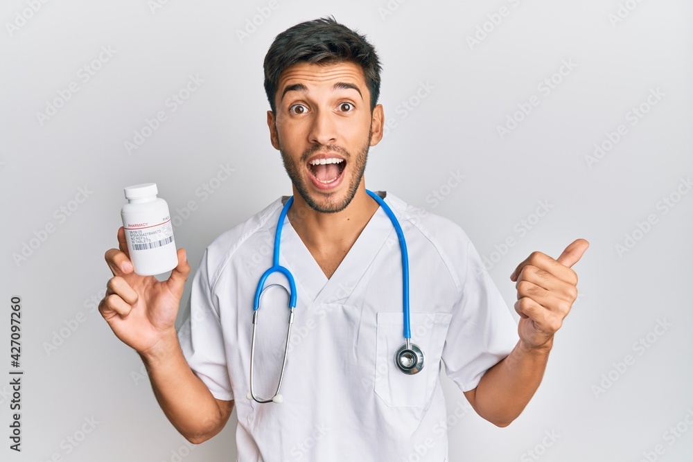 Young handsome man wearing doctor uniform holding presciption pills pointing thumb up to the side smiling happy with open mouth Young handsome man wearing doctor uniform holding presciption pills pointing thumb up to the side smiling happy with open mouth
