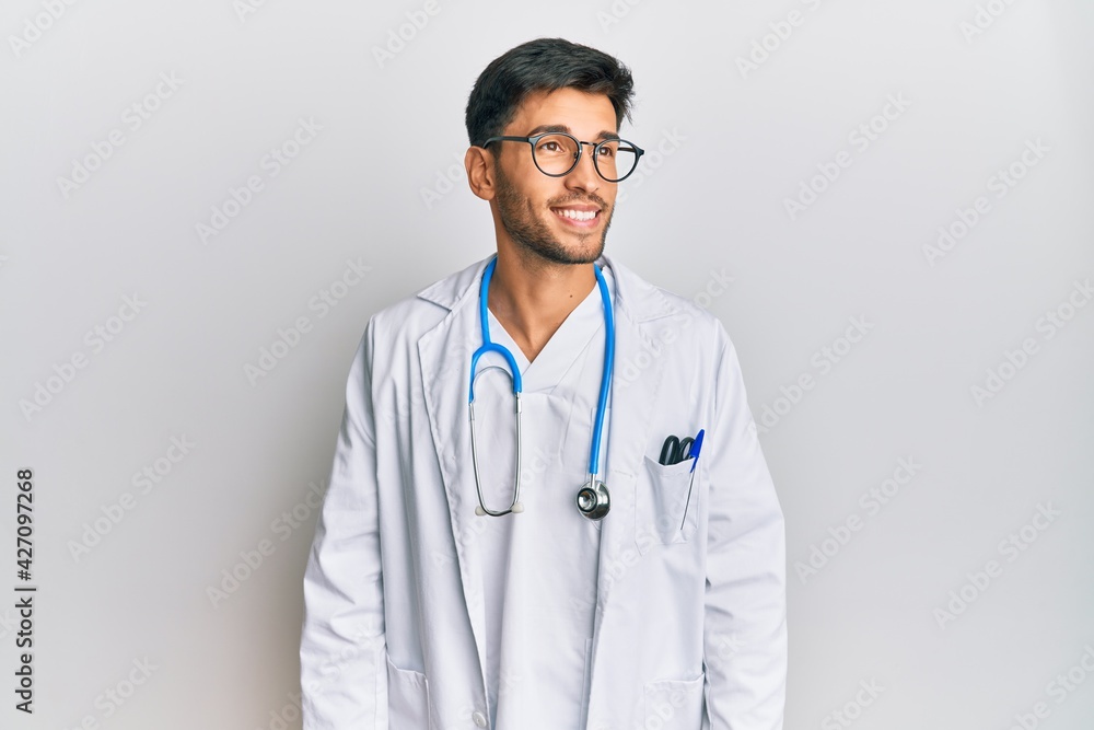 Fototapeta premium Young handsome man wearing doctor uniform and stethoscope looking away to side with smile on face, natural expression. laughing confident.