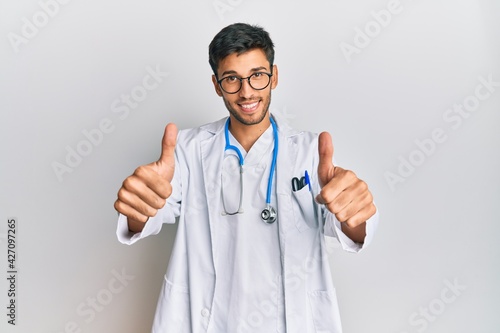 Young handsome man wearing doctor uniform and stethoscope approving doing positive gesture with hand  thumbs up smiling and happy for success. winner gesture.
