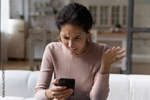 Spam mailout. Outraged nervous young latina female feel angry mad of spamming in electronic messages on smartphone. Female customer dissatisfied annoyed after reading silly email marketing letter text photo