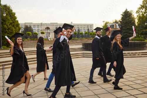 Group of young smiling university graduates in traditional mantles walking, holding diplomas in raised hands feeling happy outdoors and celebrating graduation. Successful univesity graduation concept