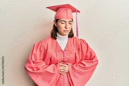 Young caucasian woman wearing graduation cap and ceremony robe with hand on stomach because indigestion, painful illness feeling unwell. ache concept.
