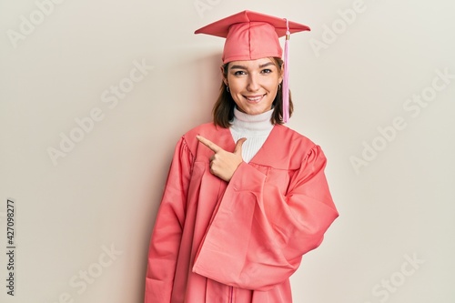 Young caucasian woman wearing graduation cap and ceremony robe cheerful with a smile of face pointing with hand and finger up to the side with happy and natural expression on face