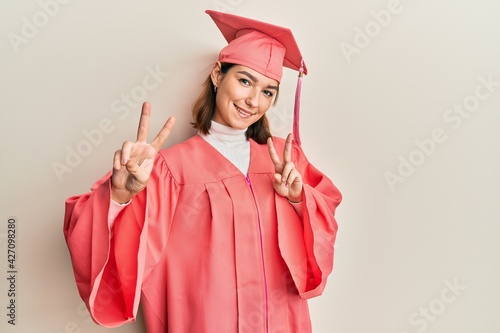 Young caucasian woman wearing graduation cap and ceremony robe smiling looking to the camera showing fingers doing victory sign. number two.