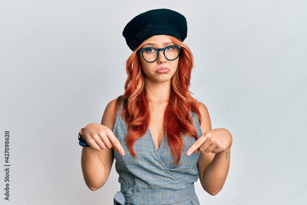 Young redhead woman wearing fashion french look with beret pointing down looking sad and upset, indicating direction with fingers, unhappy and depressed. Young redhead woman wearing fashion french look with beret pointing down looking sad and upset, indicating direction with fingers, unhappy and depressed.