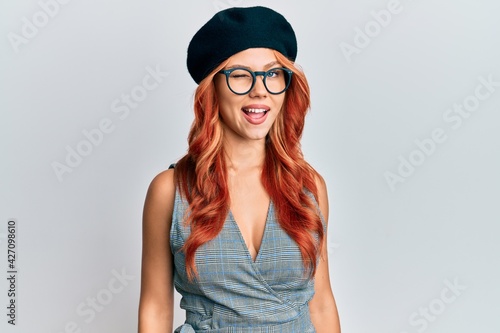 Young redhead woman wearing fashion french look with beret winking looking at the camera with sexy expression, cheerful and happy face.