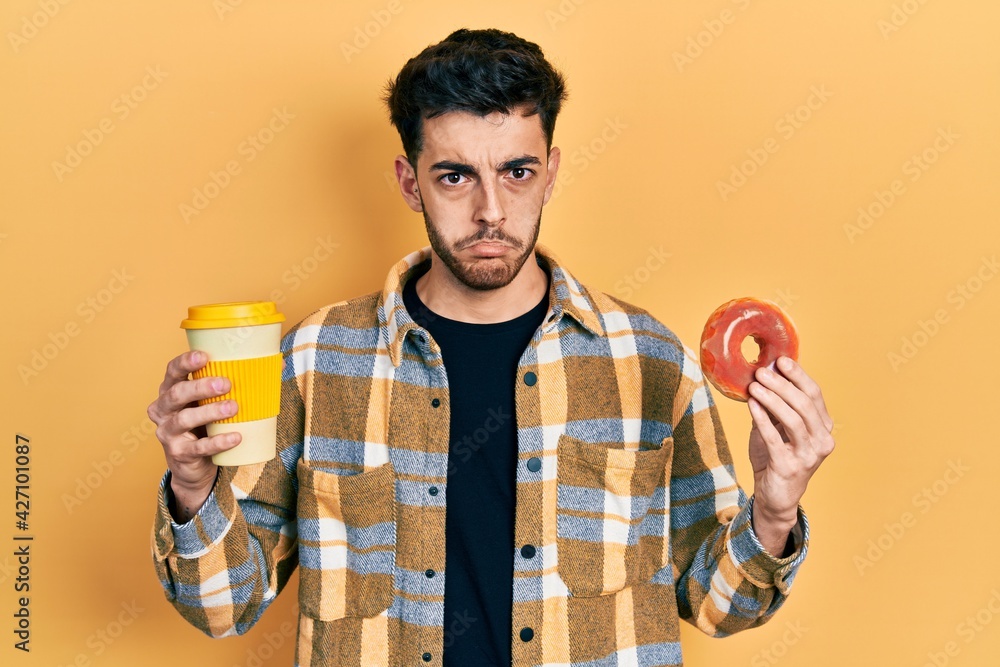 Fototapeta premium Young hispanic man eating doughnut and drinking coffee depressed and worry for distress, crying angry and afraid. sad expression.