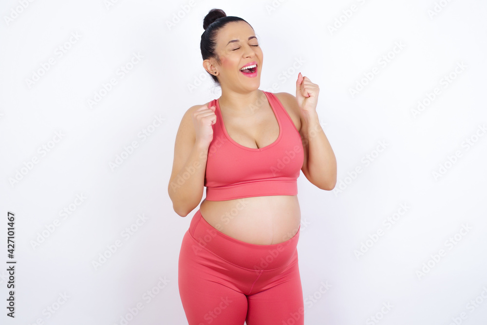 young beautiful pregnant woman in sports clothes against white wall rejoicing his success and victory clenching fists with joy being happy to achieve aim and goals. Positive emotions, feelings. young beautiful pregnant woman in sports clothes against white wall rejoicing his success and victory clenching fists with joy being happy to achieve aim and goals. Positive emotions, feelings.