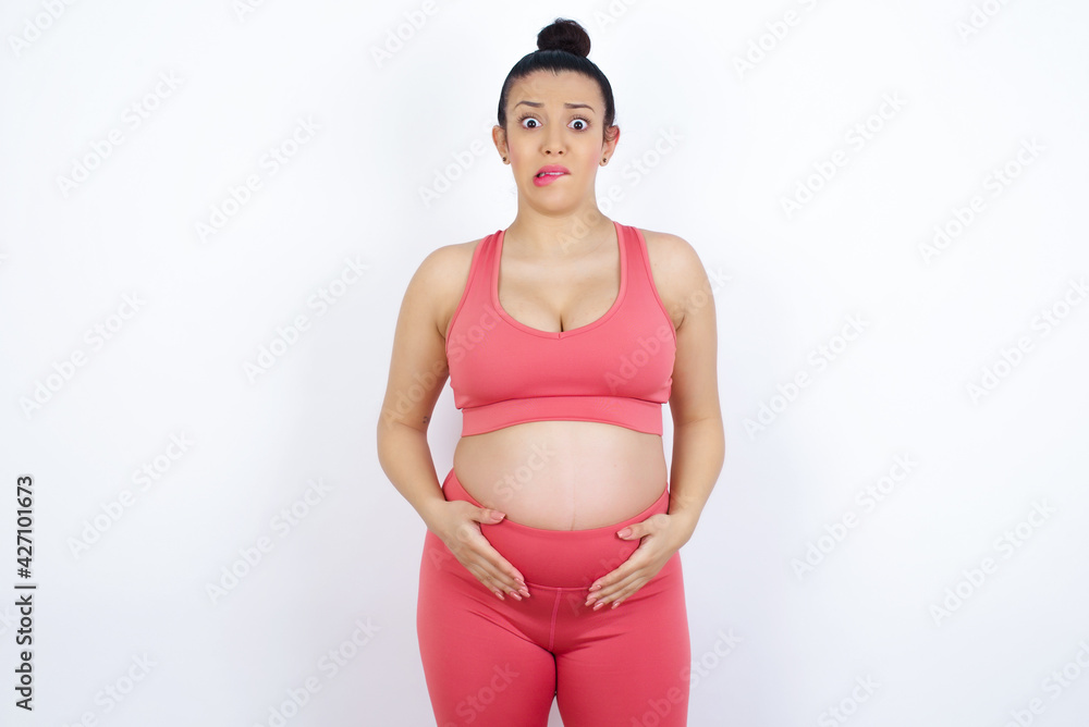 Fototapeta premium young beautiful Arab pregnant woman in sports clothes against white wall being nervous and scared biting lips looking camera with impatient expression, pensive.
