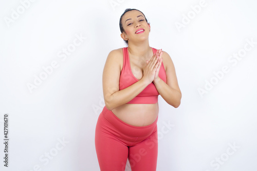 young beautiful Arab pregnant woman in sports clothes against white wall, feeling happy, smiling and clapping hands, saying congratulations with an applause.