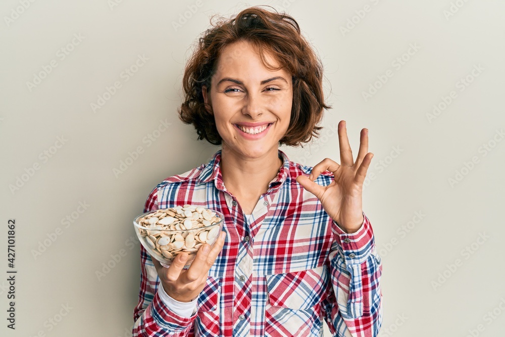 Young brunette woman holding bowl with pumpkin seeds doing ok sign with fingers, smiling friendly gesturing excellent symbol Young brunette woman holding bowl with pumpkin seeds doing ok sign with fingers, smiling friendly gesturing excellent symbol