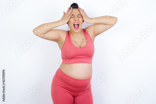 Shocked panic young beautiful Arab pregnant woman in sports clothes against white wall holding hands on head and screaming in despair and frustration. © Jihan