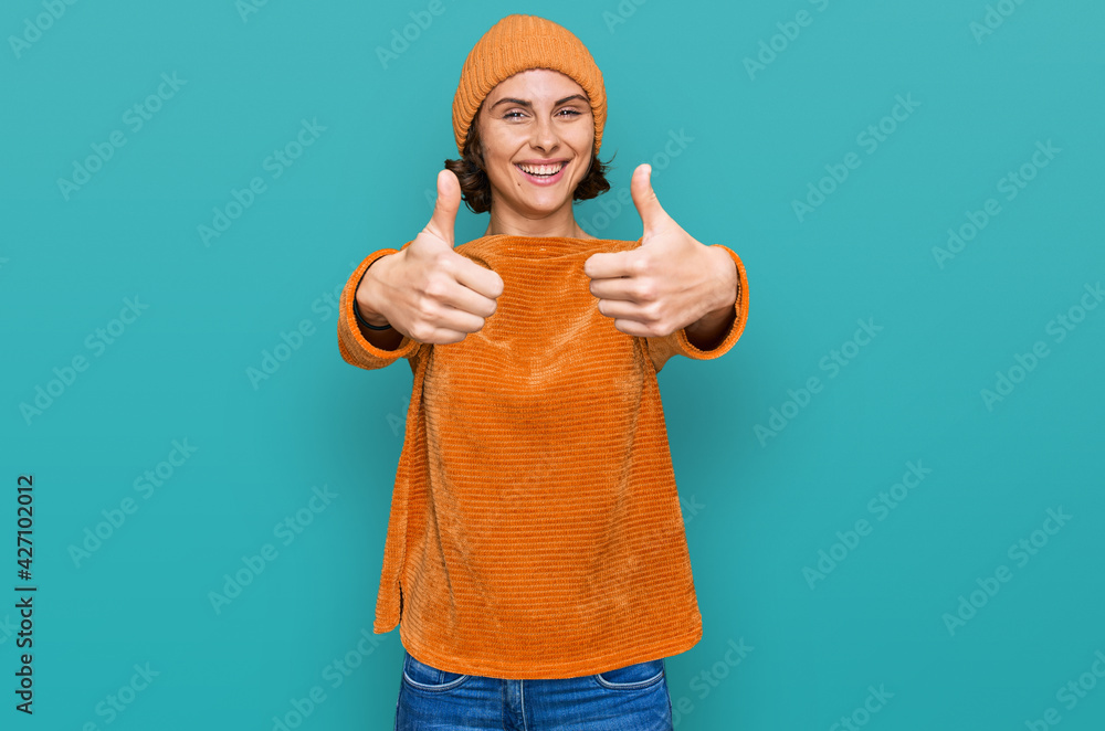 Fototapeta premium Young hispanic woman wearing casual clothes and wool cap approving doing positive gesture with hand, thumbs up smiling and happy for success. winner gesture.