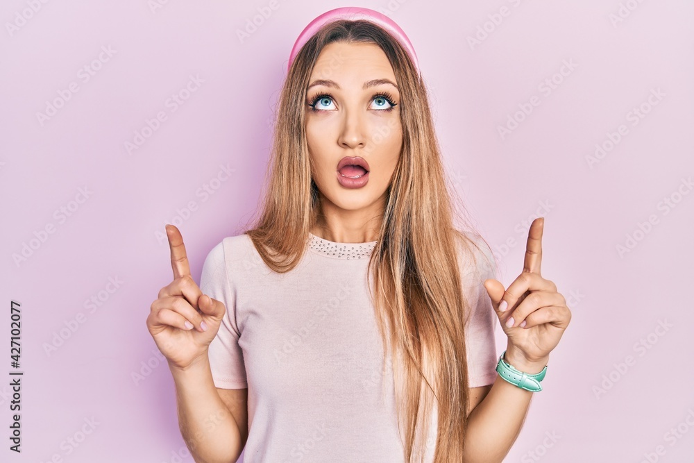 Young blonde girl wearing casual clothes amazed and surprised looking up and pointing with fingers and raised arms. Young blonde girl wearing casual clothes amazed and surprised looking up and pointing with fingers and raised arms.