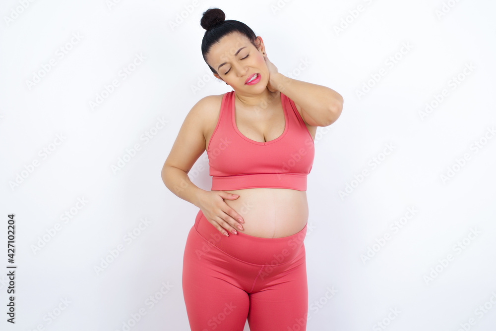 young beautiful Arab pregnant woman in sports clothes against white wall suffering from back and neck ache injury, touching neck with hand, muscular pain. young beautiful Arab pregnant woman in sports clothes against white wall suffering from back and neck ache injury, touching neck with hand, muscular pain.