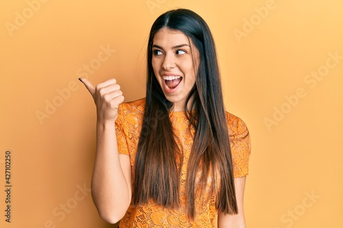 Young hispanic woman wearing casual clothes pointing thumb up to the side smiling happy with open mouth