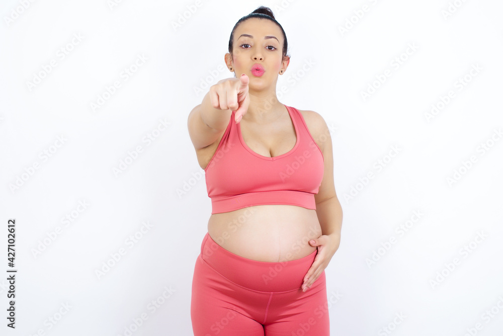 Cheerful young beautiful Arab pregnant woman in sports clothes against white wall indicates happily at you, chooses to compete, has positive expression, makes choice. Cheerful young beautiful Arab pregnant woman in sports clothes against white wall indicates happily at you, chooses to compete, has positive expression, makes choice.