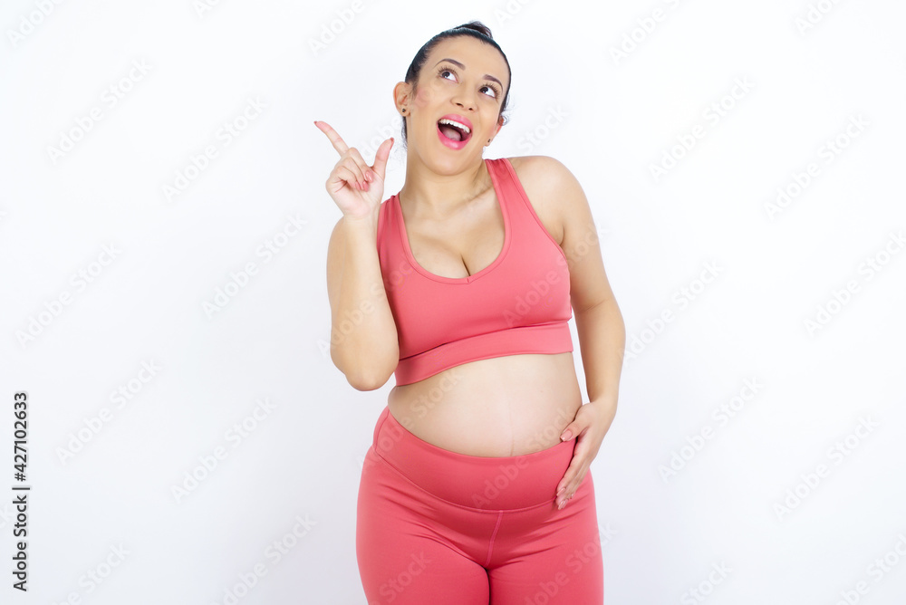 Pleasant looking young beautiful Arab pregnant woman in sports clothes against white wall has clever expression, raises one finger, remembers himself not to forget tell important thing. Pleasant looking young beautiful Arab pregnant woman in sports clothes against white wall has clever expression, raises one finger, remembers himself not to forget tell important thing.