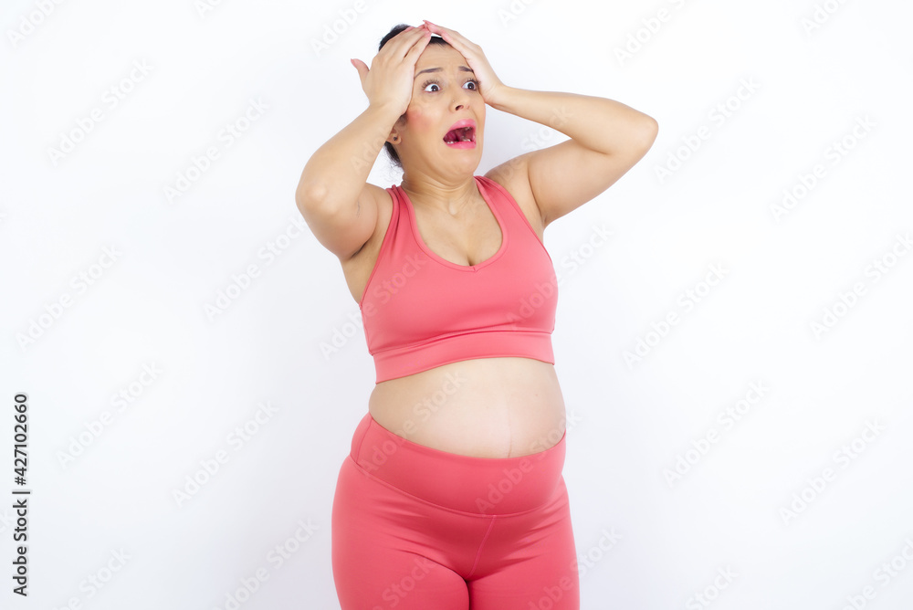 Horrible, stress, shock. Portrait emotional crazy young beautiful Arab pregnant woman in sports clothes against white wall clasping head in hands. Emotions, facial expression concept. Horrible, stress, shock. Portrait emotional crazy young beautiful Arab pregnant woman in sports clothes against white wall clasping head in hands. Emotions, facial expression concept.
