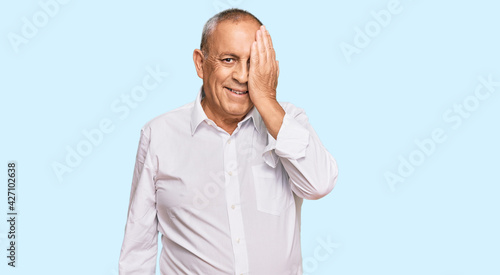 Handsome senior man wearing elegant white shirt covering one eye with hand, confident smile on face and surprise emotion. © Krakenimages.com