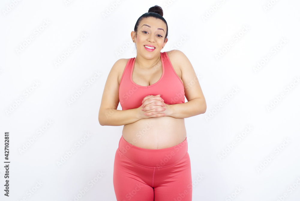 Happy young beautiful Arab pregnant woman in sports clothes against white wall stands against orange studio wall keeps hands on heart, swears be loyal, expresses gratitude. Honesty concept. Happy young beautiful Arab pregnant woman in sports clothes against white wall stands against orange studio wall keeps hands on heart, swears be loyal, expresses gratitude. Honesty concept.