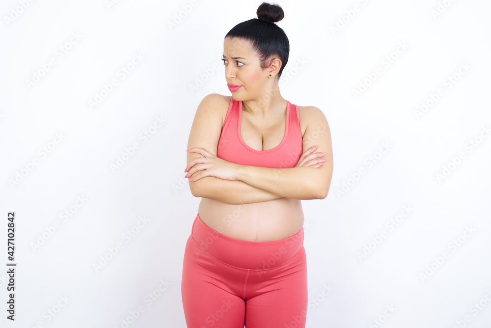 Displeased young beautiful Arab pregnant woman in sports clothes against white wall with bad attitude, arms crossed looking sideways. Negative human emotion facial expression feelings. Displeased young beautiful Arab pregnant woman in sports clothes against white wall with bad attitude, arms crossed looking sideways. Negative human emotion facial expression feelings.