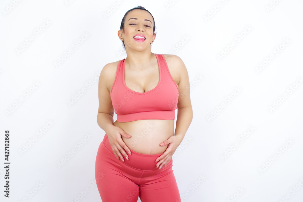 young beautiful Arab pregnant woman in sports clothes against white wall with happy and funny face smiling and showing tongue. young beautiful Arab pregnant woman in sports clothes against white wall with happy and funny face smiling and showing tongue.