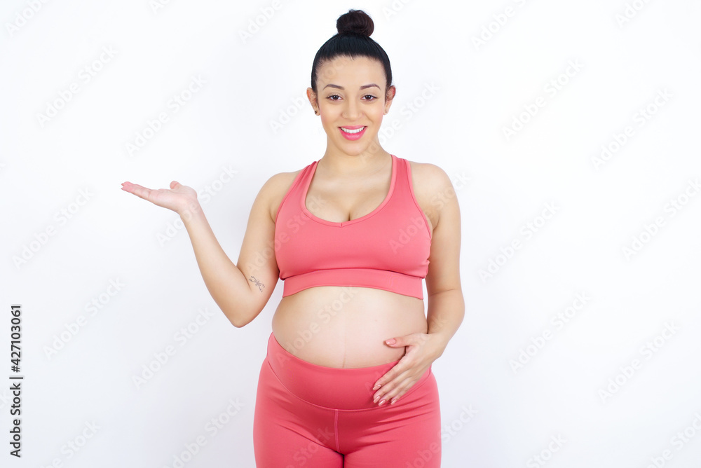Positive glad young beautiful Arab pregnant woman in sports clothes says: wow how exciting it is, has amazed expression, shows something on blank space with open hand. Advertisement concept. Positive glad young beautiful Arab pregnant woman in sports clothes says: wow how exciting it is, has amazed expression, shows something on blank space with open hand. Advertisement concept.