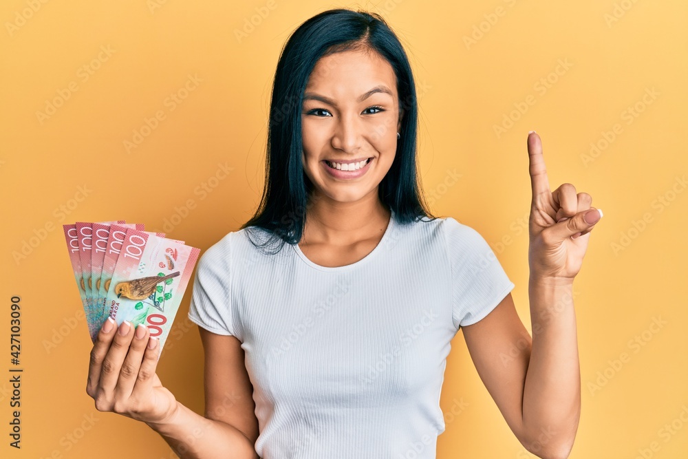 Beautiful hispanic woman holding 100 new zealand dollars banknote smiling with an idea or question pointing finger with happy face, number one Beautiful hispanic woman holding 100 new zealand dollars banknote smiling with an idea or question pointing finger with happy face, number one