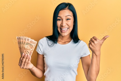 Beautiful hispanic woman holding 50 brazilian real banknotes pointing thumb up to the side smiling happy with open mouth