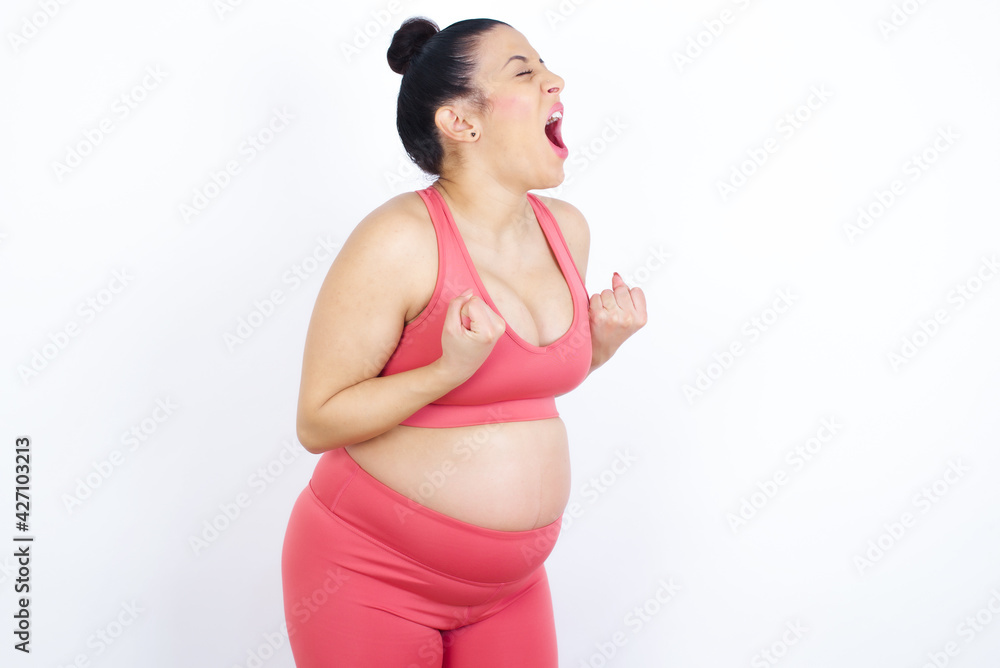 young beautiful Arab pregnant woman in sports clothes against white wall excited and glad to achieve victory, clenches fists, screams in excitement with closed eyes,successful person. young beautiful Arab pregnant woman in sports clothes against white wall excited and glad to achieve victory, clenches fists, screams in excitement with closed eyes,successful person.