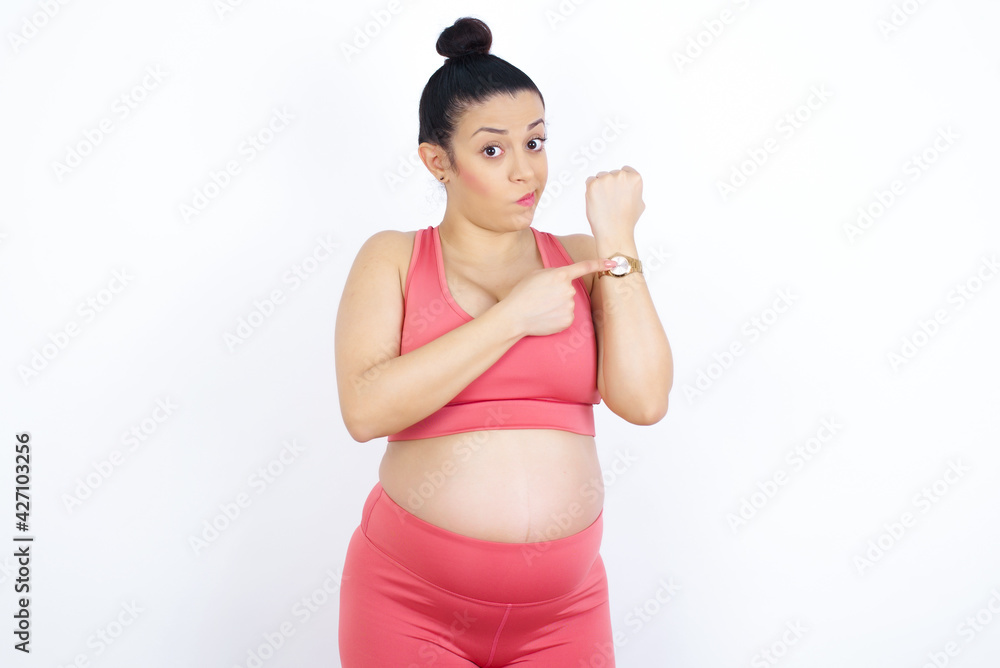 young beautiful Arab pregnant woman in sports clothes against white wall in hurry pointing to watch time, impatience, upset and angry for deadline delay young beautiful Arab pregnant woman in sports clothes against white wall in hurry pointing to watch time, impatience, upset and angry for deadline delay