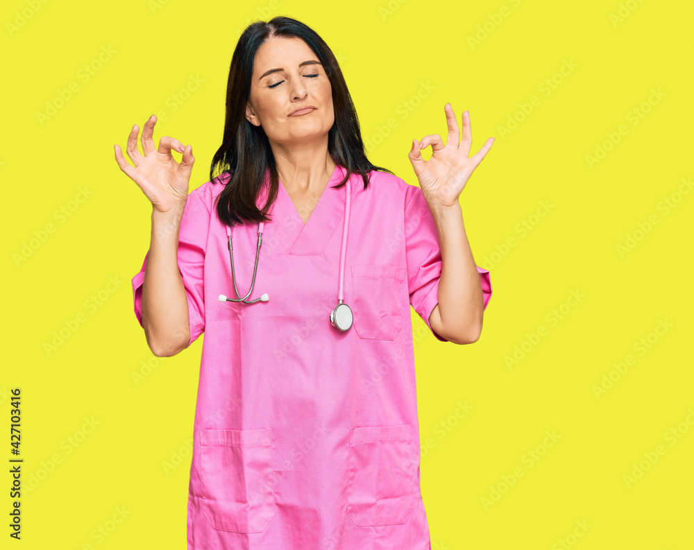 Middle age brunette woman wearing doctor uniform and stethoscope relax and smiling with eyes closed doing meditation gesture with fingers. yoga concept. Middle age brunette woman wearing doctor uniform and stethoscope relax and smiling with eyes closed doing meditation gesture with fingers. yoga concept.